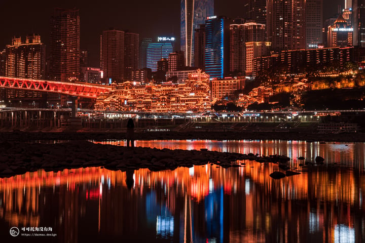 A nighttime cityscape is reflected in calm water, with tall buildings illuminated by lights and a bridge crossing the river.