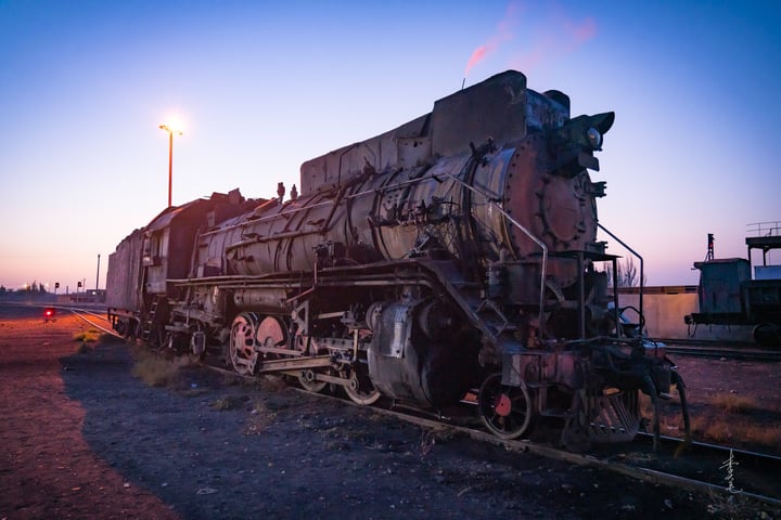 An old, black steam locomotive with a white front and black body is parked on a track, emitting smoke from its chimney. The train is surrounded by a serene landscape of a blue sky and a tall streetlight.