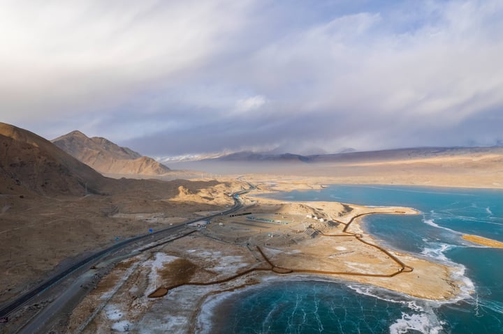 A serene landscape features a deep blue lake reflecting the sky, with snow-capped mountains in the distance. The sky is filled with white clouds, and the landscape is dominated by brown and beige tones.