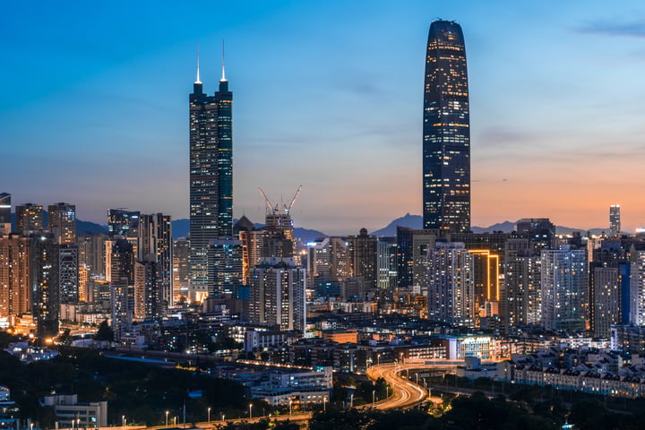 A city skyline at dusk, with a prominent skyscraper, a curved building, and a mountain range in the background.