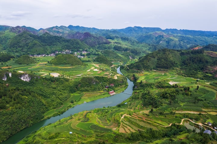 The image captures the stunning Guizbou, with lush green mountains and rice terraces in the foreground. The river meanders through the valley, surrounded by houses and buildings, and the sky is a light blue with white clouds. The perspective is from an aerial view, showcasing the harmony between nature and human habitation.