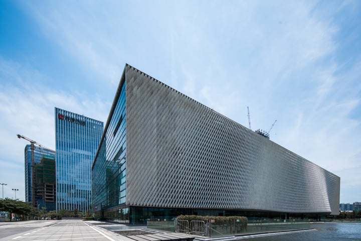 A modern, rectangular building with a geometric facade and reflective windows stands in a cityscape. It is situated on a sidewalk, with a body of water nearby. The sky is clear blue with a few clouds, and a crane is visible in the background. The building's design and location suggest it could be a museum or a public building.