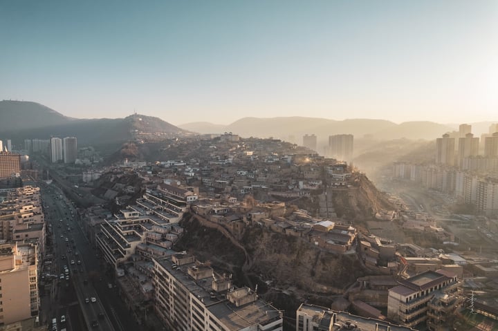 An aerial view of a mountainous city with a hillside, densely populated with buildings of varying heights, surrounded by greenery. The city is situated on a hill, with roads and highways running through it. The sun casts a warm glow on the city, highlighting its architectural details.