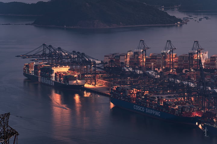A large cargo ship is docked in a harbor, surrounded by cranes and other ships, with a mountain range in the background.