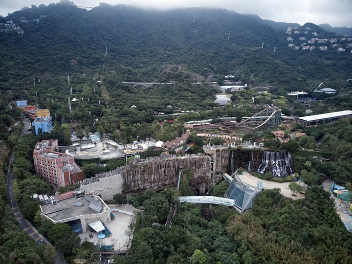 An aerial view of a mountainous area with a waterfall, surrounded by lush greenery and buildings, with a bridge connecting two parts of the landscape.