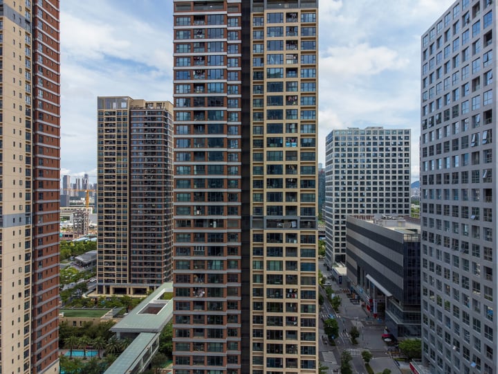 A high vantage point captures a bustling cityscape with tall buildings, a mix of brown and gray, and a clear blue sky with scattered clouds.