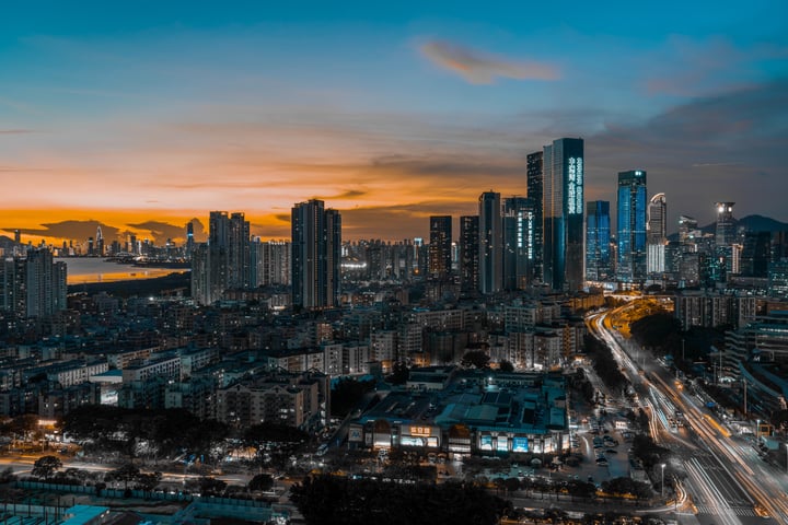A city skyline at sunset, with tall buildings and a dark blue sky, illuminated by street lights and cars, and a body of water in the distance.