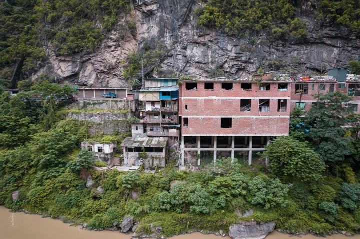 An old, multi-level brick building with a blue roof is perched on a cliff overlooking a river. The building is surrounded by lush greenery and is partially submerged in brown water.