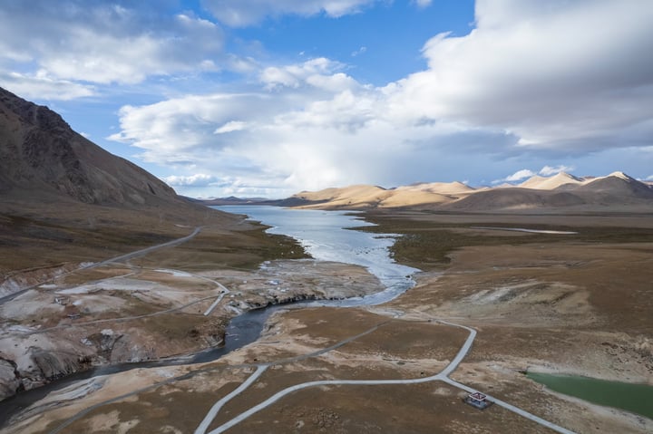 An aerial view of a mountainous landscape with a river flowing through it, surrounded by mountains and a blue sky with clouds.