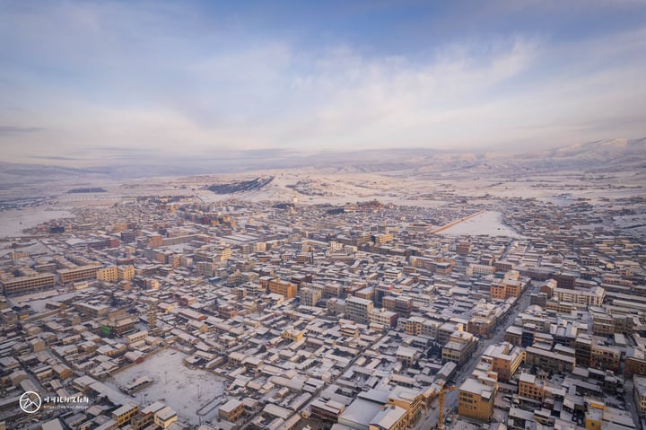 An aerial view of a snowy city with a mix of buildings and roads, surrounded by mountains and a clear blue sky.