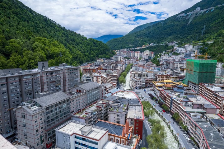 A cityscape with a river, buildings, and mountains, with a clear blue sky and green trees.