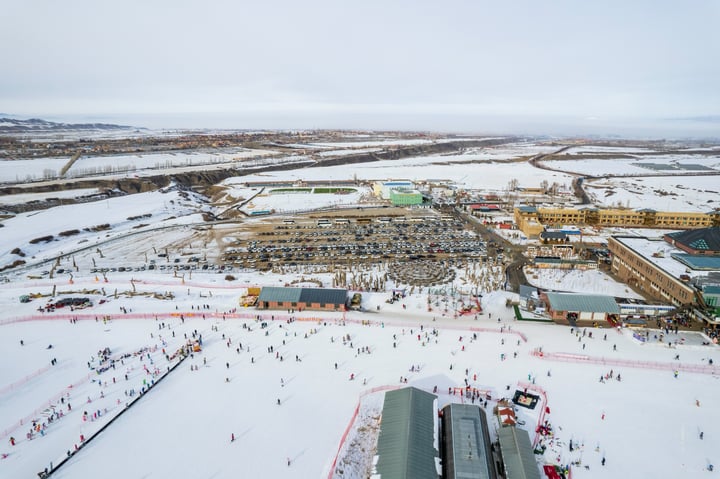 An aerial view of a snowy landscape with a large skier's area, surrounded by buildings and a parking lot, with a large sports complex in the background.