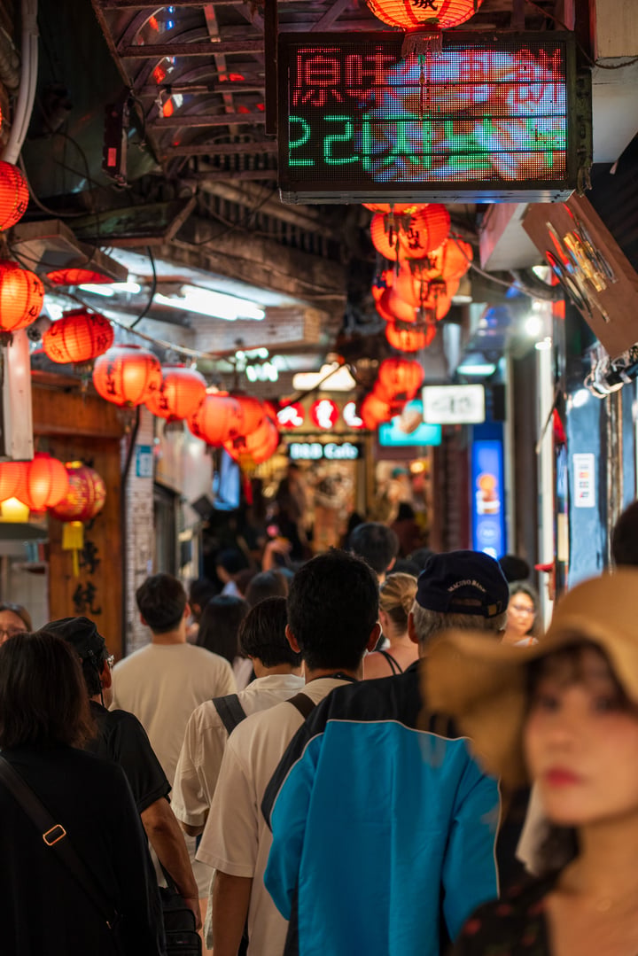 A bustling street filled with people, shops, and lanterns, with a man in a hat walking down the street.
