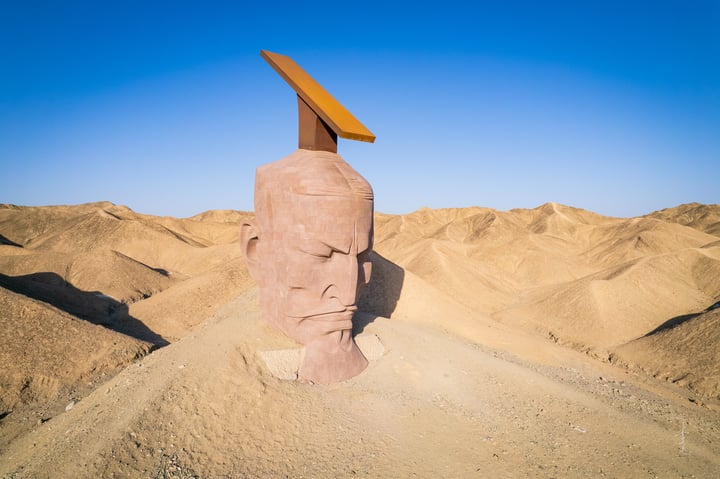 A large, pinkish-brown sand sculpture of a face with a yellow sun on top, standing on a hill in a desert landscape.