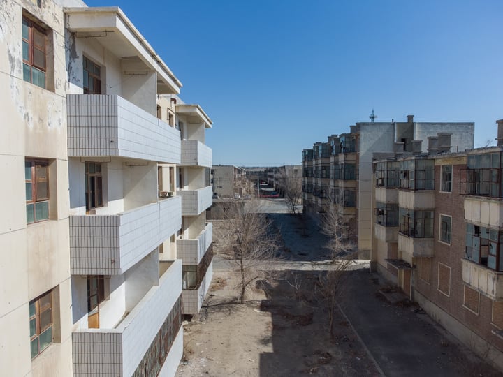 A cityscape with three white apartment buildings, one with a balcony, surrounded by a dirt road and trees.