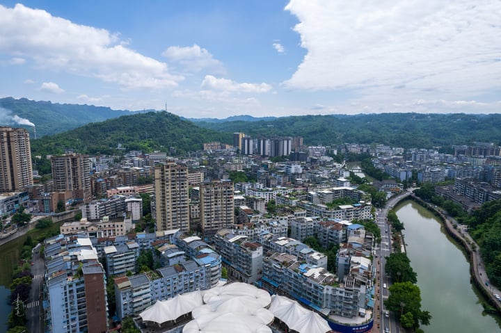 An aerial view of a city nestled between mountains, with tall buildings and a large white dome in the center, surrounded by lush greenery.