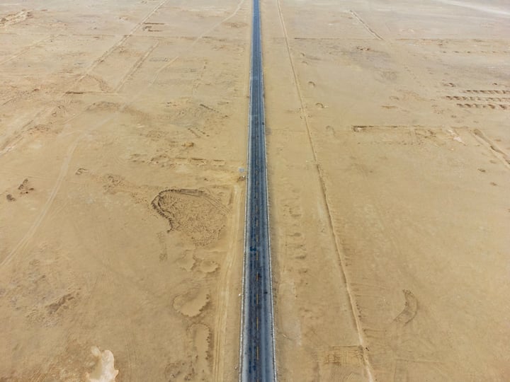An aerial view of a sandy landscape with a long, straight, dark blue line running through it, surrounded by footprints and tracks.