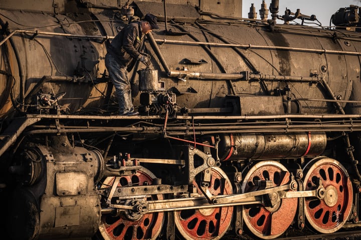 An old black steam locomotive with red wheels is traveling on a railway track, emitting smoke from its chimney.