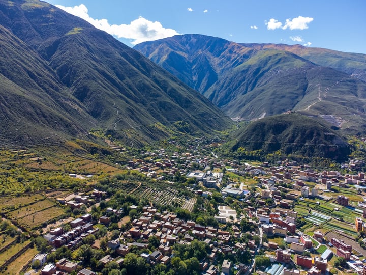 A serene valley with a small town nestled between mountains, surrounded by lush greenery and a clear blue sky.