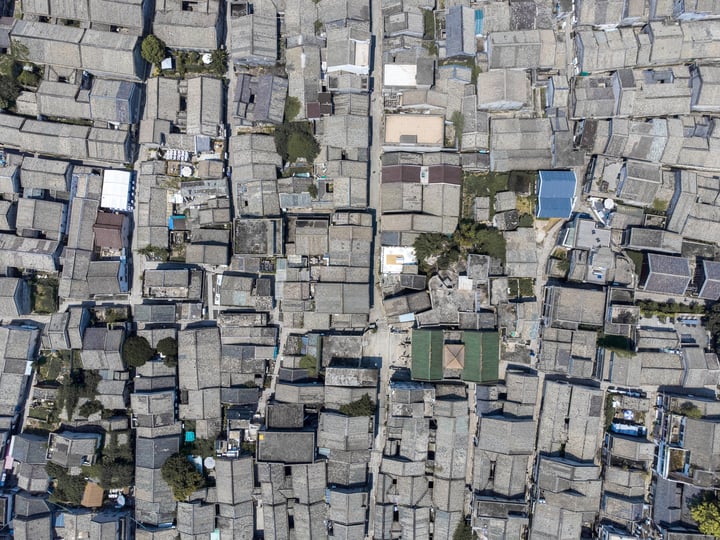 An aerial view of a densely populated city with a mix of gray and brown buildings, some with green roofs, surrounded by trees and green spaces.