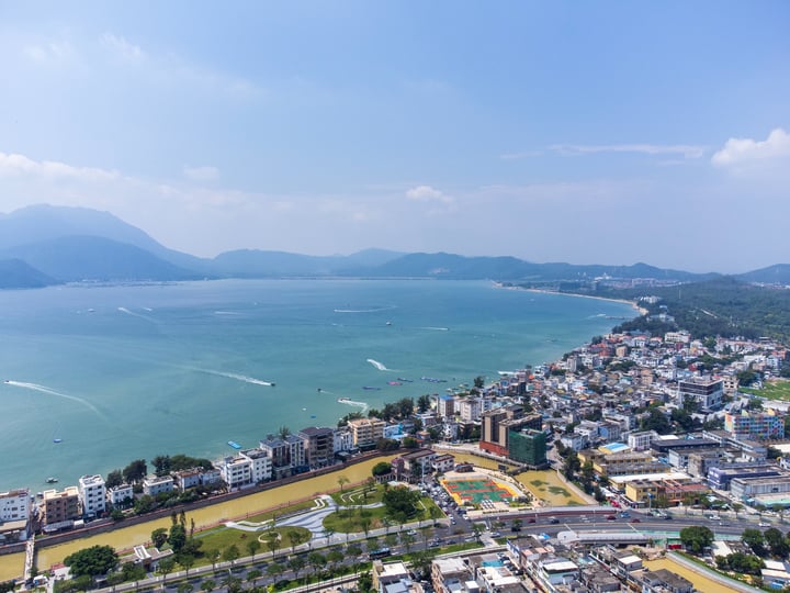 A panoramic view of a coastal city with a large body of water, boats, and mountains in the background.