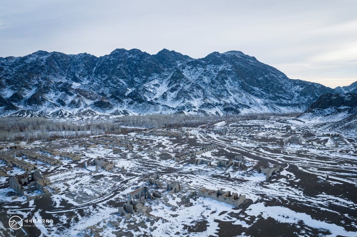 A snowy mountain range with snow-covered peaks and valleys, bare trees, and a small village in the foreground.