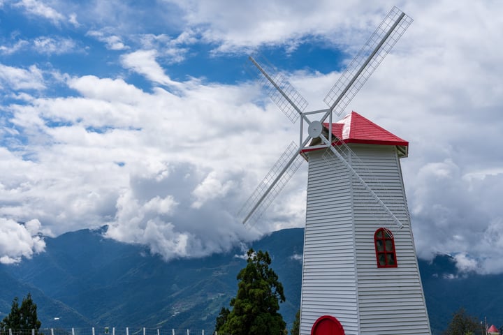 A white windmill with a red roof stands tall in a lush green landscape, surrounded by trees and mountains. The windmill has four white sails and a red window, and is set against a backdrop of a blue sky with white clouds.
