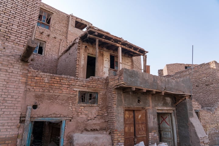 An old, dilapidated brick building with a wooden door and balcony, surrounded by debris and a pile of bricks, stands in a desert-like environment.