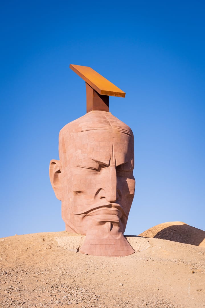 A large, pinkish-brown sculpture of a man's head with a large, orange, pointed object on top, set against a clear blue sky.