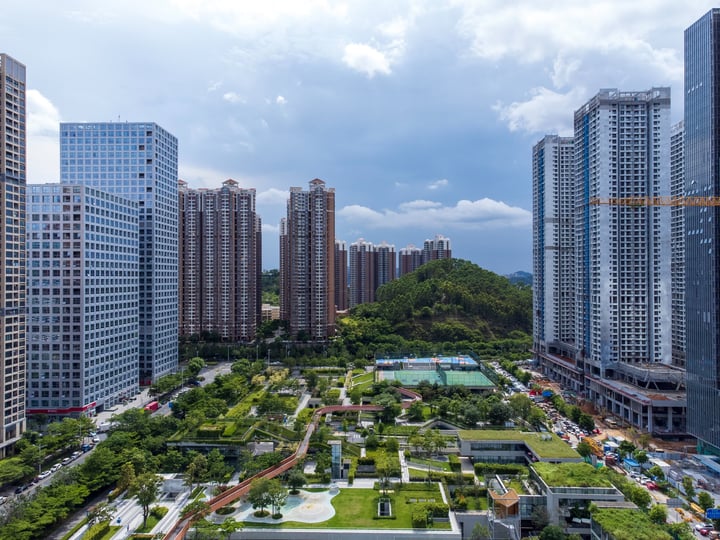 A cityscape with tall buildings, a park, and a basketball court, with a clear blue sky and clouds.