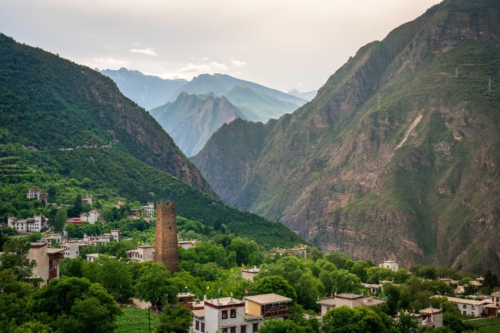 A serene mountainous valley with a quaint village, surrounded by lush greenery and towering mountains, is captured in a high-angle view. The village is nestled amidst verdant hills, and the sky is a soft light blue with a few clouds. The image is taken from a higher vantage point, offering a panoramic view of the valley and mountains.