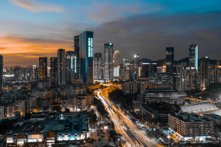 A city skyline at dusk, with tall buildings illuminated by street lights, a highway with moving cars, and a vibrant sky.
