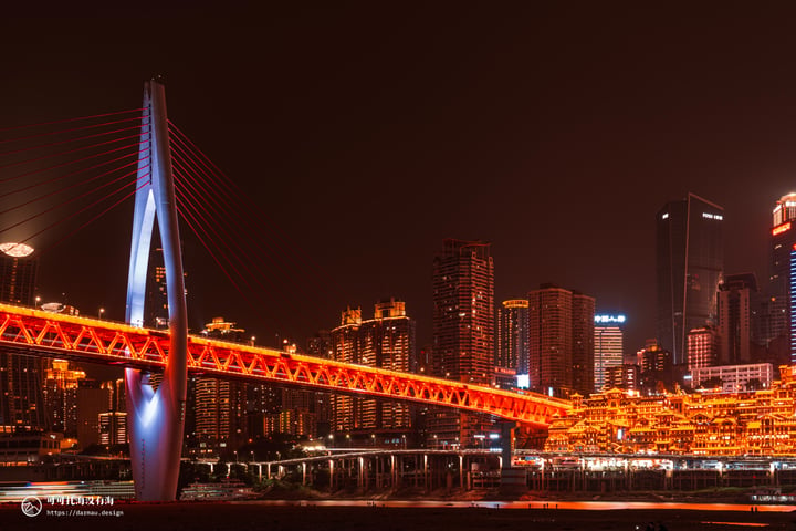 A nighttime cityscape features a red suspension bridge with white pillars, illuminated by orange lights, spanning a body of water. Tall buildings with lit windows and a "Mars" logo are in the background, and a dark sky contrasts the vibrant cityscape.