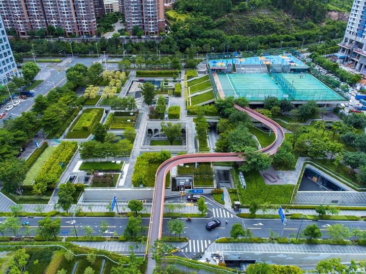 An aerial view of a city park with a curved road, lush greenery, and a tennis court, surrounded by tall buildings.