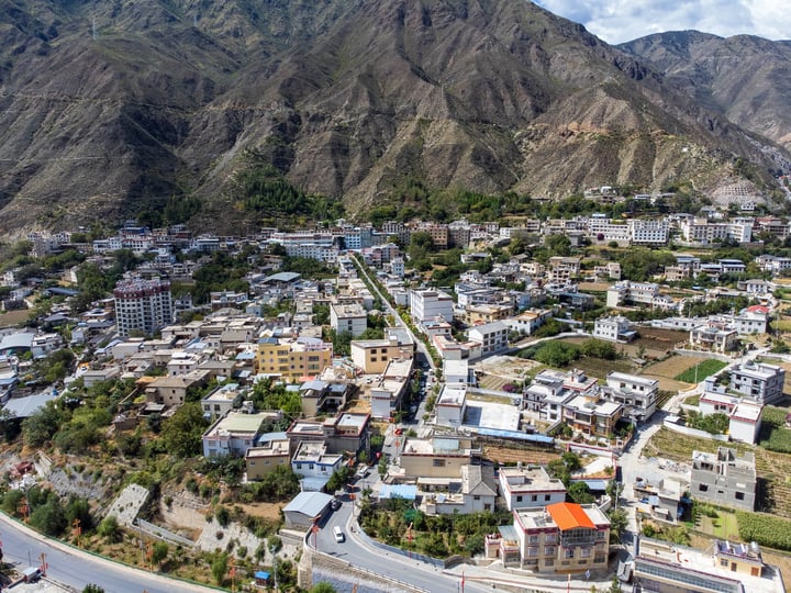 A serene mountain town with white and gray buildings, orange roofs, and green vegetation, surrounded by a winding road and a clear blue sky.