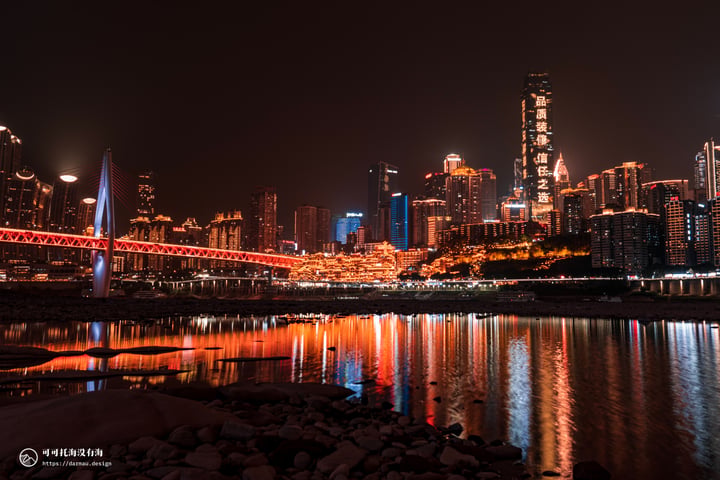 A nighttime cityscape is reflected in calm water, with tall buildings illuminated by lights and a bridge crossing the river.