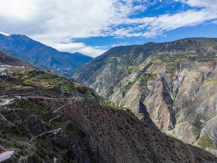 A winding mountain road winds through a valley, surrounded by steep cliffs and lush greenery, with a clear blue sky above.