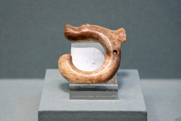 A carved wooden fish sculpture is displayed on a gray stand, with a white stone plaque beneath it. The fish has a curved body and a pointed head, and is placed on a gray pedestal. The background is a blue wall.