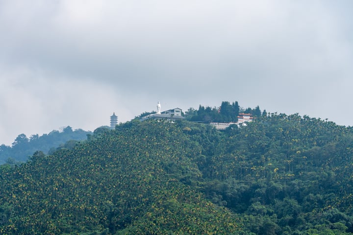 A white building with a red roof sits atop a hill, surrounded by lush greenery and a cloudy sky.