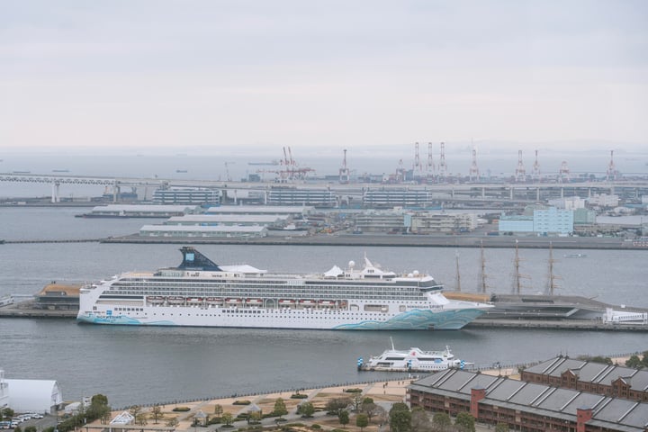A large white cruise ship with a blue stripe is docked in a harbor, surrounded by smaller boats and cranes. The harbor is bustling with activity, and the city skyline is visible in the background.