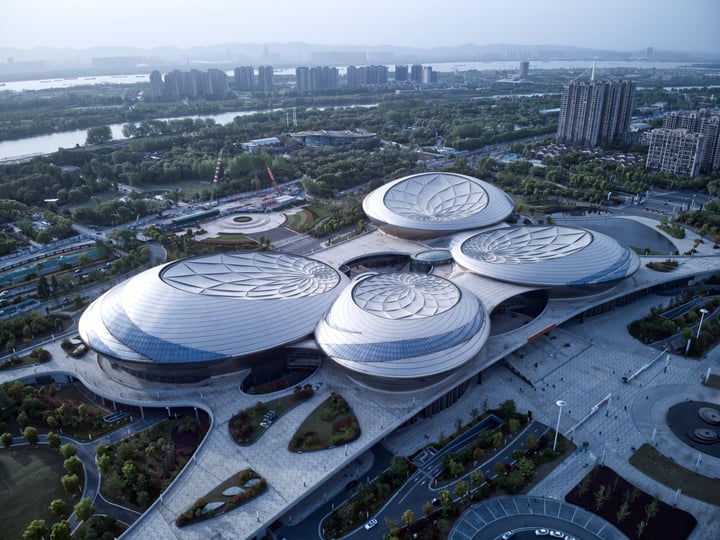 An aerial view of a modern, futuristic building complex with three circular structures, surrounded by a park and a river, with a city skyline in the background.
