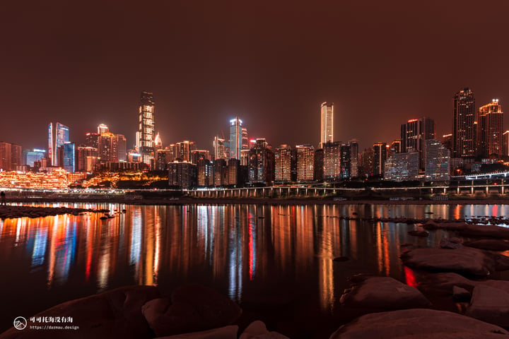 A nighttime cityscape is reflected in calm water, with tall buildings illuminated by lights and a bridge crossing the river.