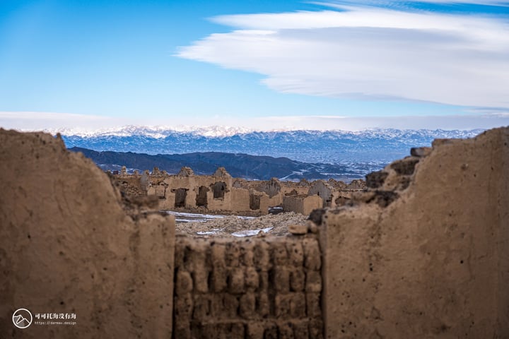 The image features a mountain range with snow-capped peaks, a vast sky with white clouds, and a weathered, brown brick building in the foreground. The perspective is from a high vantage point, looking down on the ruins.