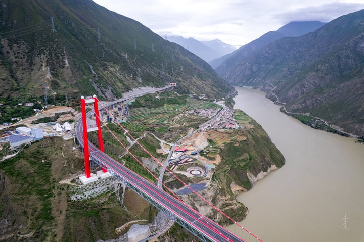A red suspension bridge spans a river in a valley, with a white building on the left and a red bridge on the right. The river is calm, and the sky is cloudy.