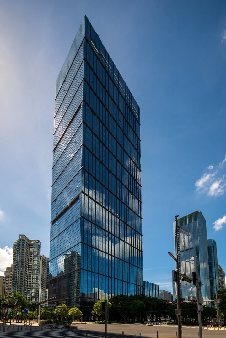 A modern skyscraper with blue glass facade stands tall in a bustling cityscape, reflecting the sky and surrounding buildings. The building's design is sleek and contemporary, with a triangular top and a flat bottom. The city street in front of it is lined with trees and streetlights, adding a touch of nature to the urban environment. The image captures the harmony between architecture and nature in a modern city.