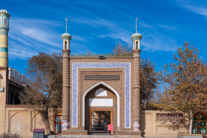 A serene scene features a large, ornate archway with blue and white tiles, flanked by two green minarets and a blue dome. The archway is set against a backdrop of a clear blue sky with scattered clouds. The archway is surrounded by lush green trees and a fence, creating a tranquil atmosphere.