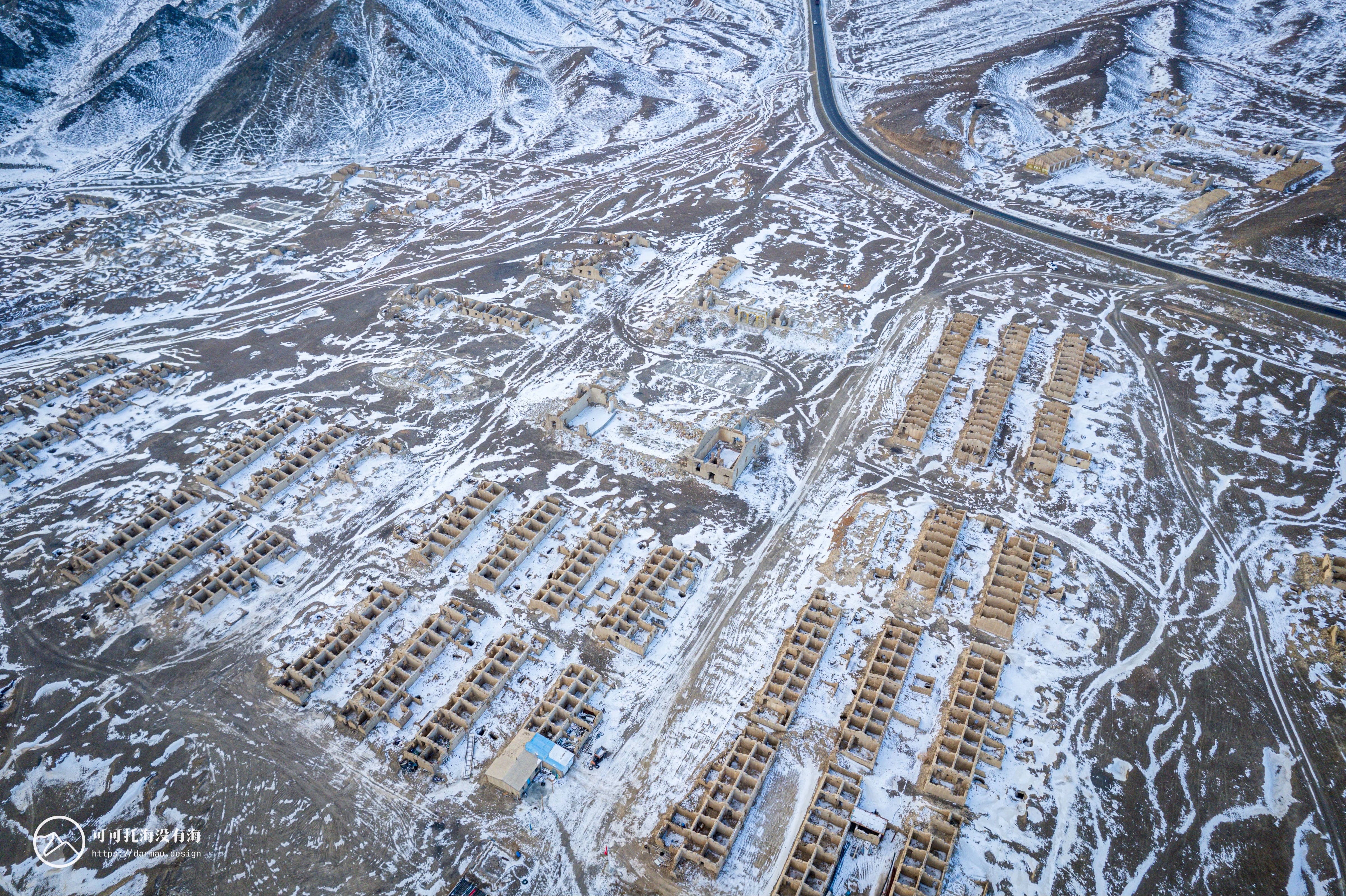 An aerial view of a snowy landscape with a road running through it, surrounded by wooden buildings and a mountain range.