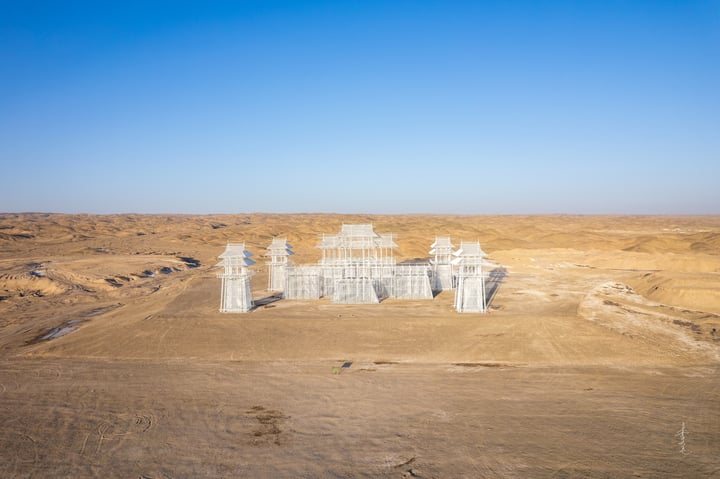 An aerial view of a white, pagoda-like structure with multiple levels and a central roof, surrounded by a barren desert landscape.