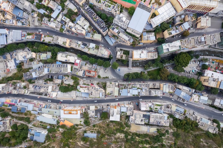 An aerial view of a city with a complex network of roads, including a winding road, surrounded by buildings of varying heights and colors. The city is densely populated with cars and buses, and the roads are well-lit. The buildings are predominantly white and gray, with some featuring orange roofs. The city is surrounded by greenery, and the sky is visible in the top left corner.