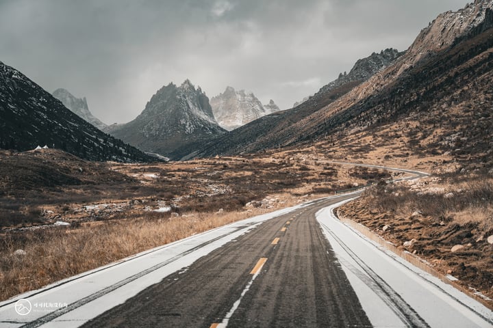 A winding, black asphalt road with tire tracks cuts through a mountainous landscape, surrounded by patches of grass and snow. The sky is filled with clouds, casting a soft light over the scene.